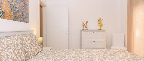 A bedroom with a patterned bedspread, a white dresser, a nightstand, a wall-mounted air conditioner, and a painting on the wall.