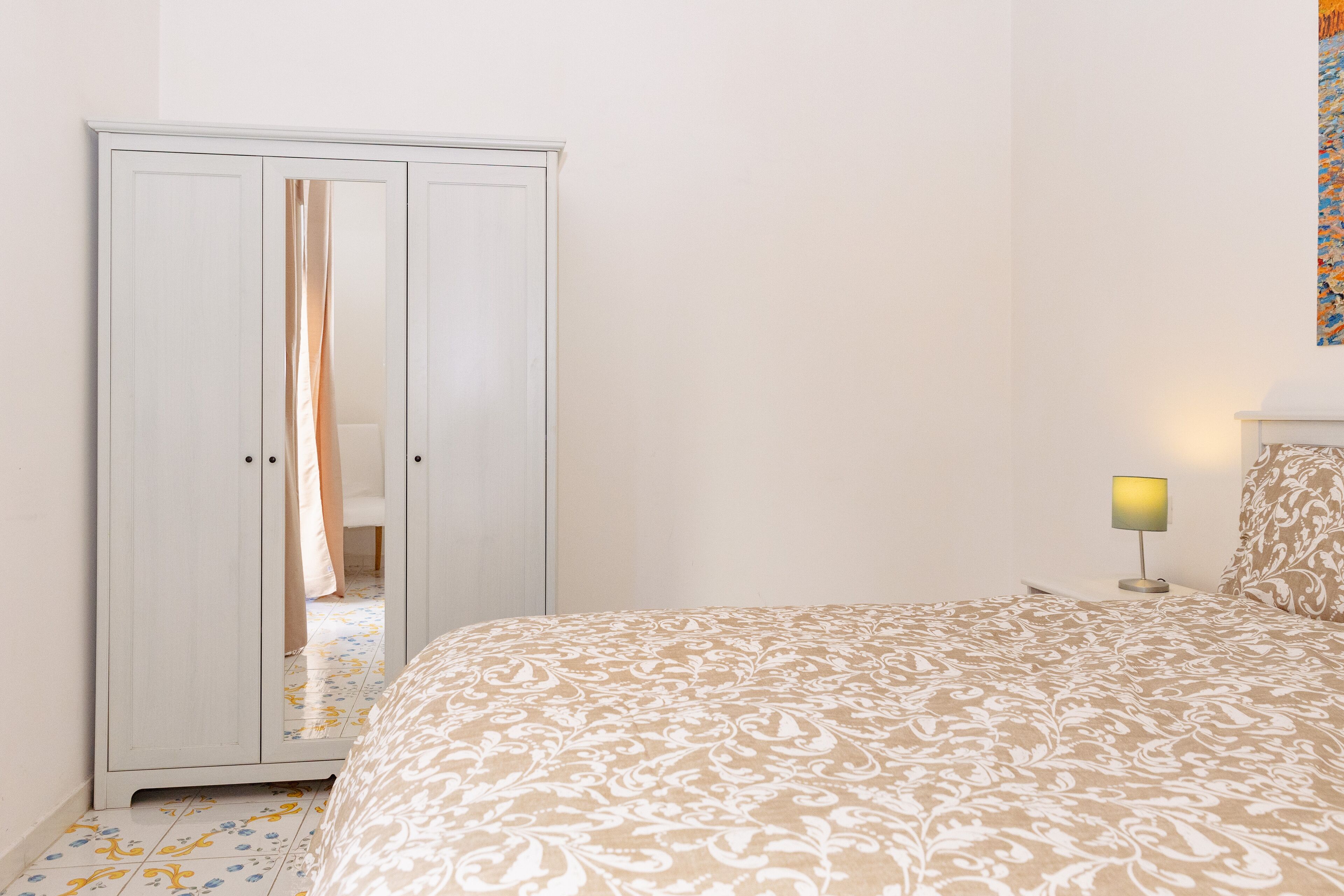 A bedroom with a bed, a wardrobe, a nightstand with a lamp, and a wall with a painting.