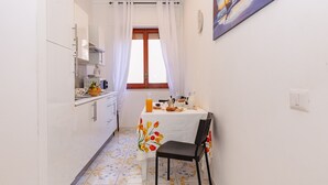 A narrow kitchen with white cabinets, a dining table with a floral tablecloth, and a chair. A window with curtains is visible, and a painting hangs on the wall. - Residence Sorrento (Sorrento)