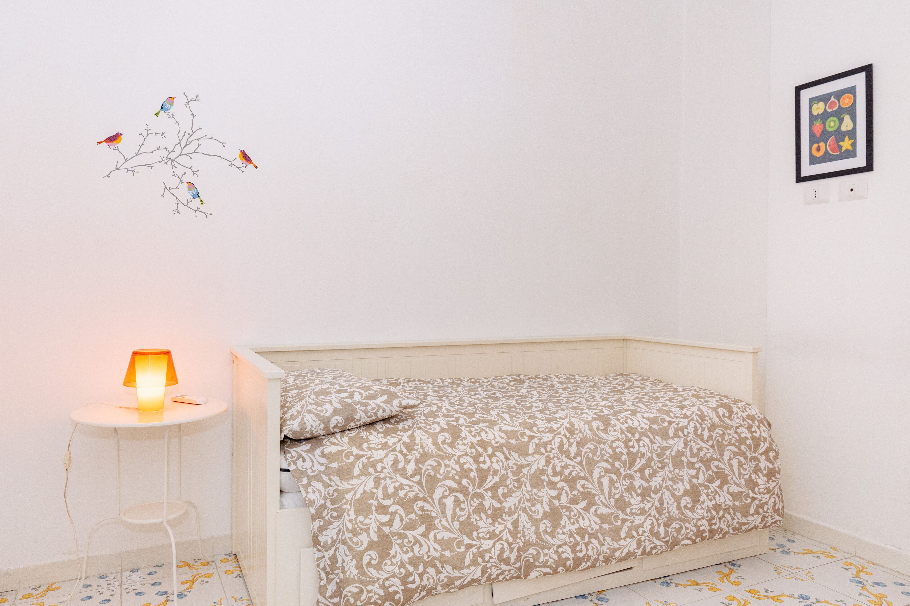 Quarto com cama, mesa de cabeceira com abajur, arte na parede com pássaros e um quadro com frutas e legumes.