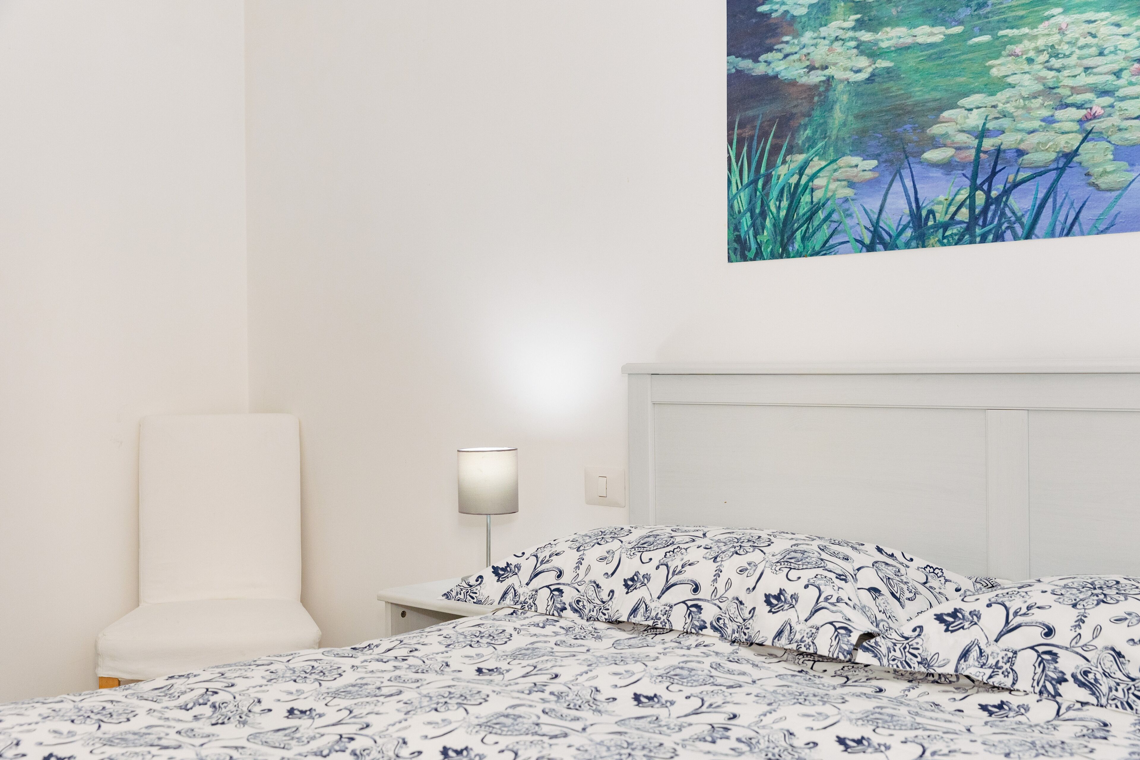 A bedroom with a bed, a nightstand, a lamp, and a wall-mounted painting of flowers.