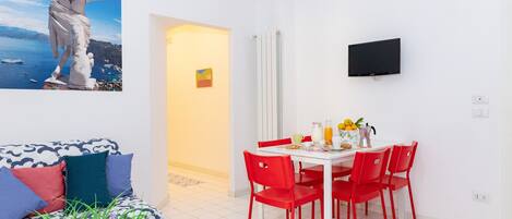 A modern living room with a white sofa, red chairs, a dining table, and a TV mounted on the wall.