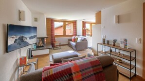 Ett modernt vardagsrum med en soffa, ett soffbord och en tv som är monterad på väggen. Det finns ett fönster med gardiner, ett litet bord med en lampa och en hylla med dekorativa föremål.