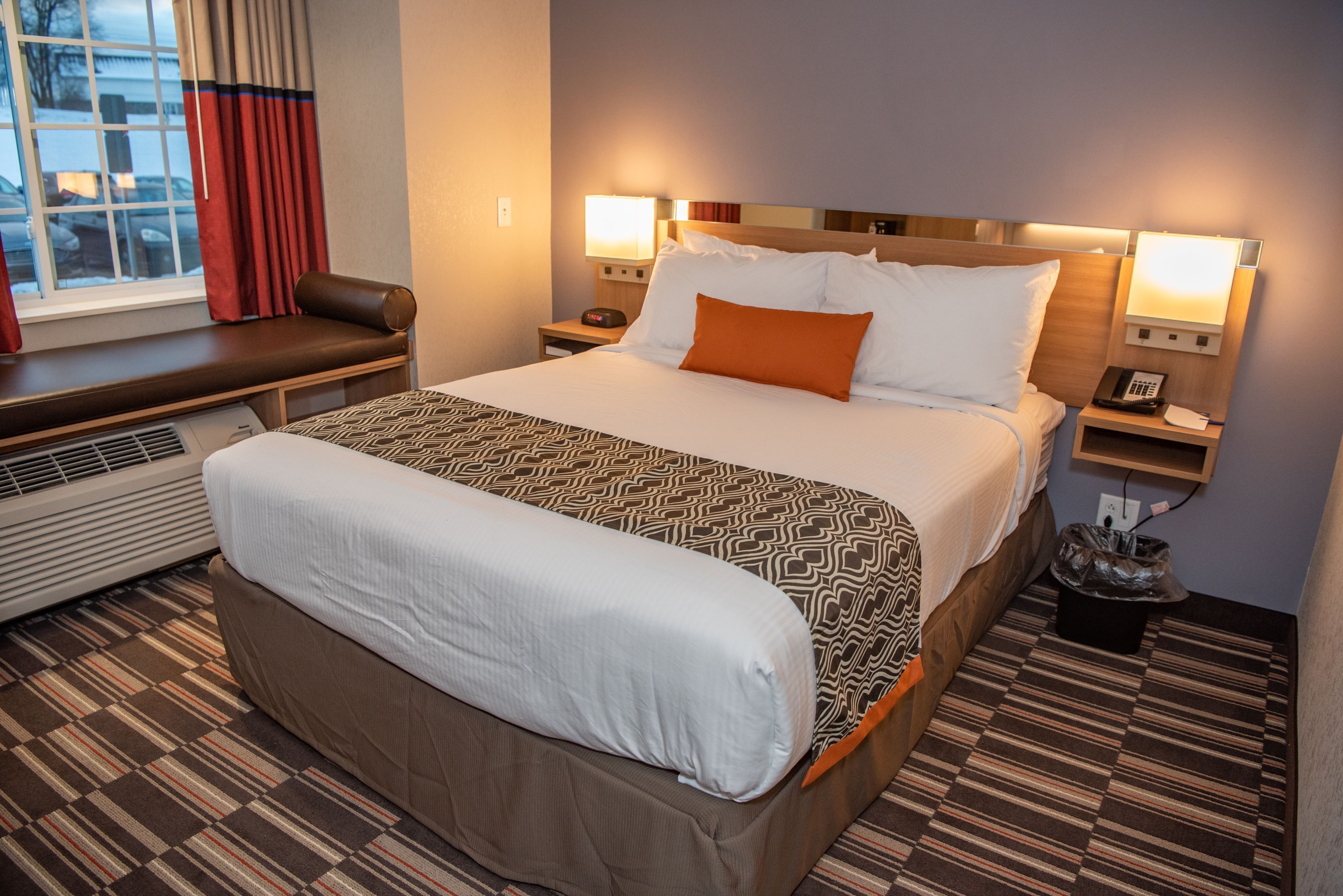 Habitación de hotel con una cama grande, un banco junto a la ventana, un calentador y un cesto de basura.