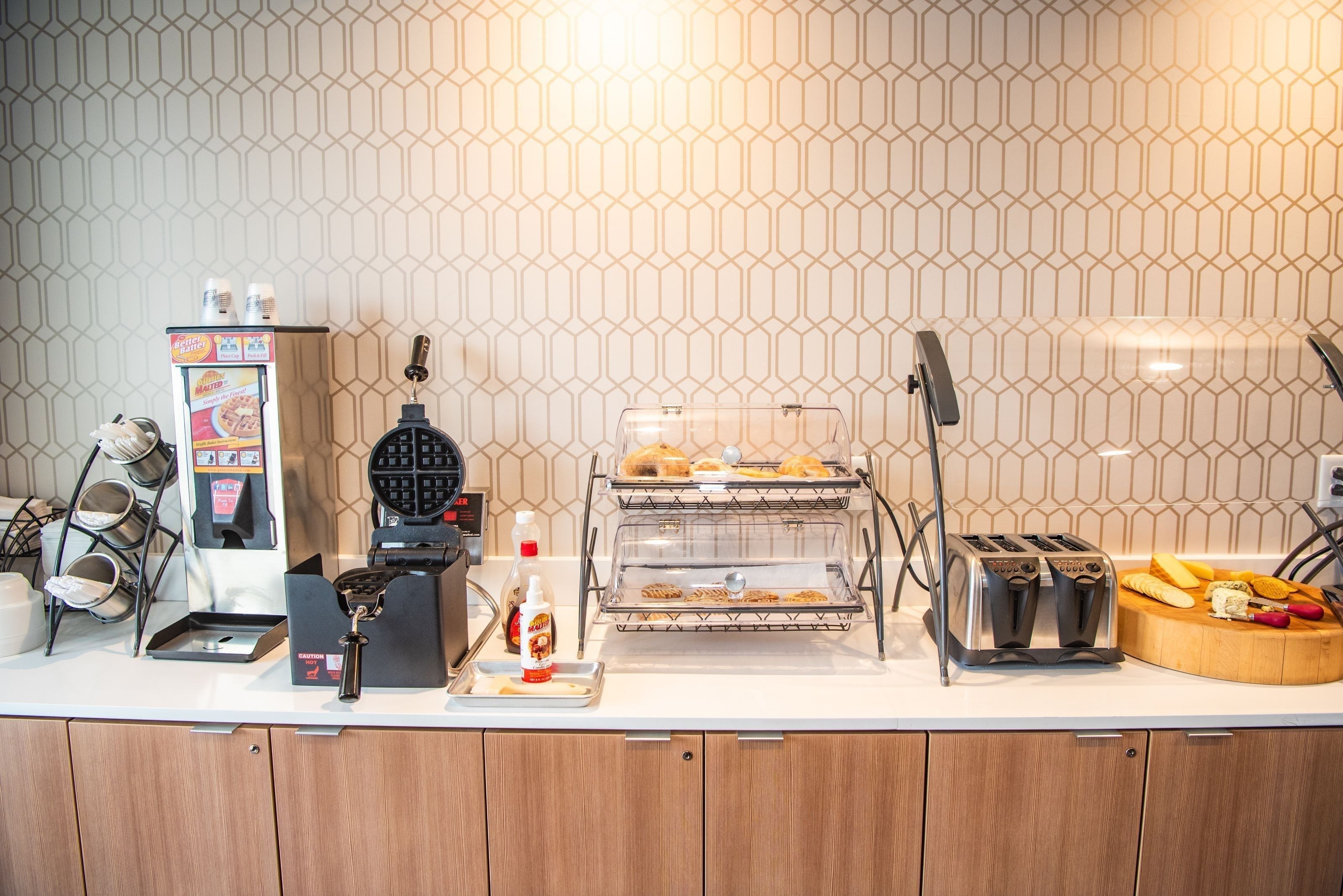 A breakfast bar with waffle maker, toaster, and a display case with pastries.