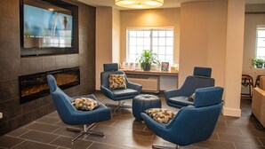 A modern lounge area with a flat-screen TV mounted on the wall, a fireplace, and several blue armchairs with patterned cushions. - Microtel Inn & Suites by Wyndham Carlisle (Carlisle)
