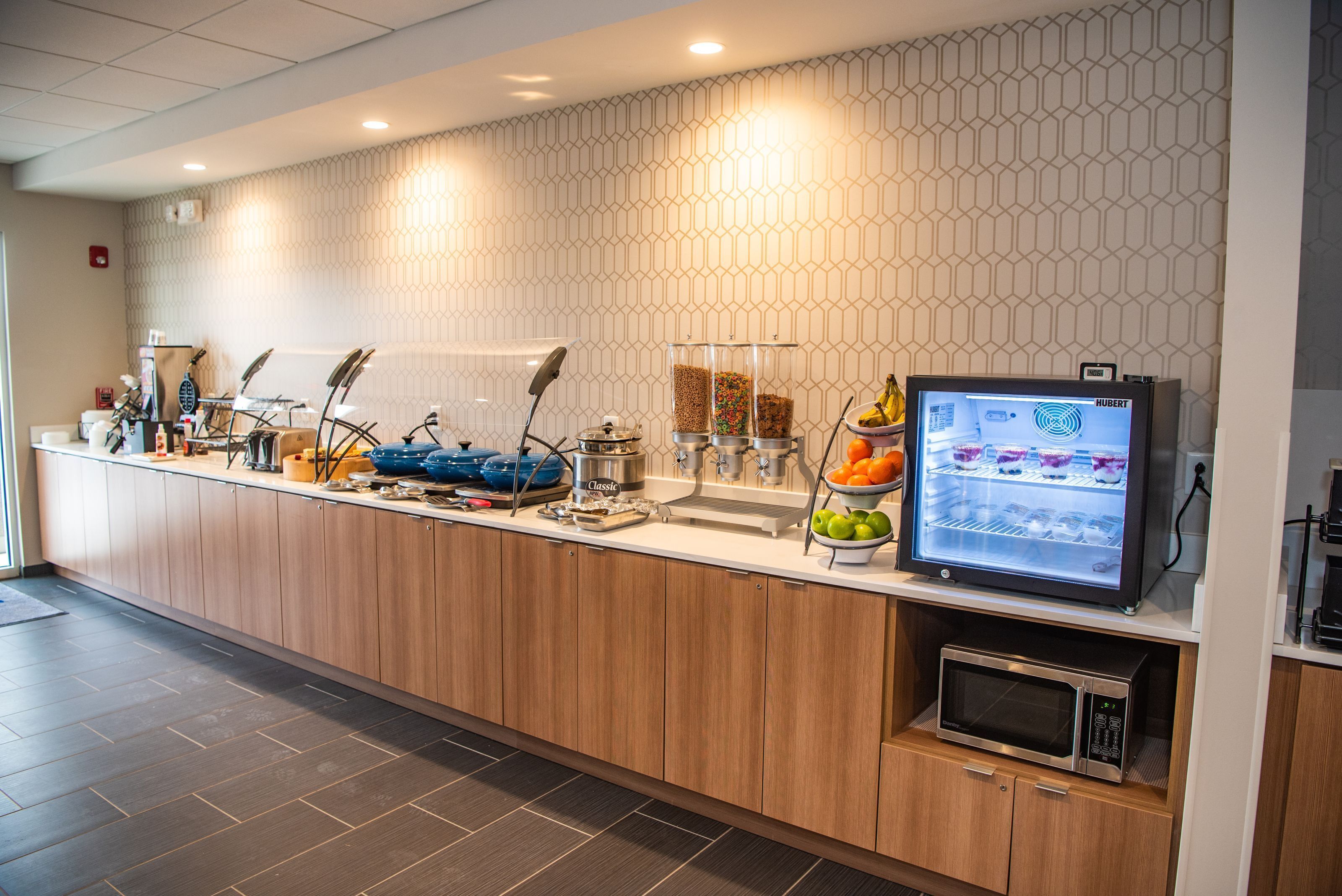 A modern hotel reception area with a wooden counter, a coffee station, a fruit bowl, a microwave, and a digital screen displaying the hotel's name.