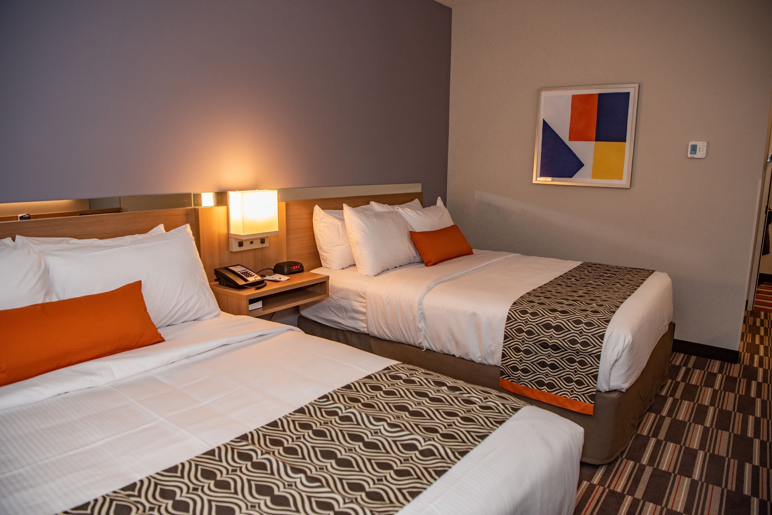 Ein Hotelzimmer mit zwei Betten, jeweils mit gemusterten Bettbezügen, einem Nachttisch mit Lampe und einem an der Wand befestigten Kunstwerk.