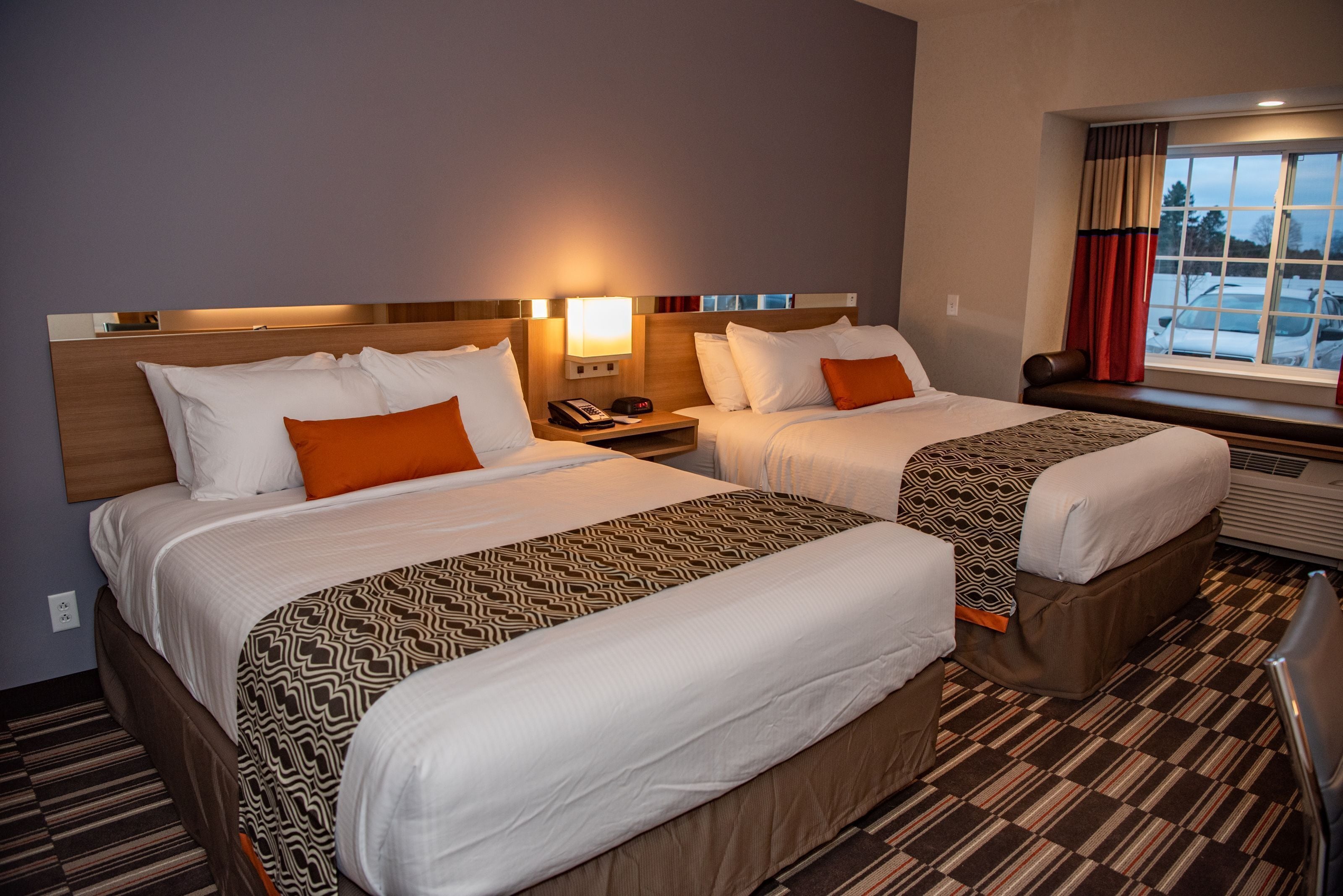 Habitación de hotel con dos camas, una mesita de noche con lámpara, ventana con cortinas y vista a coches estacionados afuera.