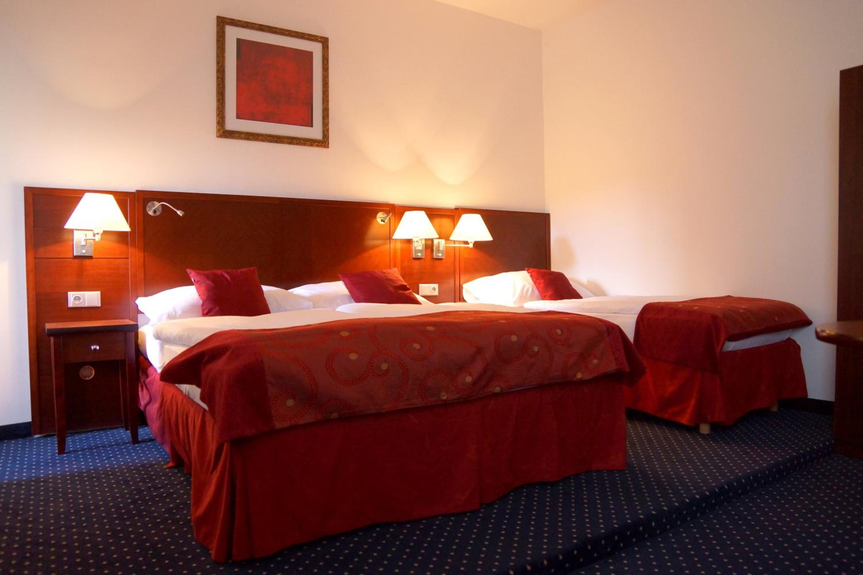 Habitación de hotel con dos camas, cada una con ropa de cama y cabeceros rojos. Hay dos lámparas de noche y un cuadro en la pared.