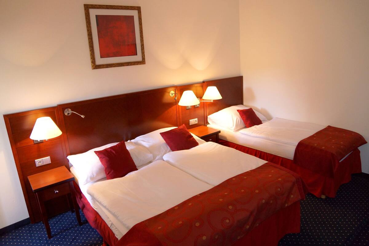Una habitación de hotel con dos camas, cada una con una colcha roja, una mesita de noche con una lámpara y un cuadro colgado en la pared.