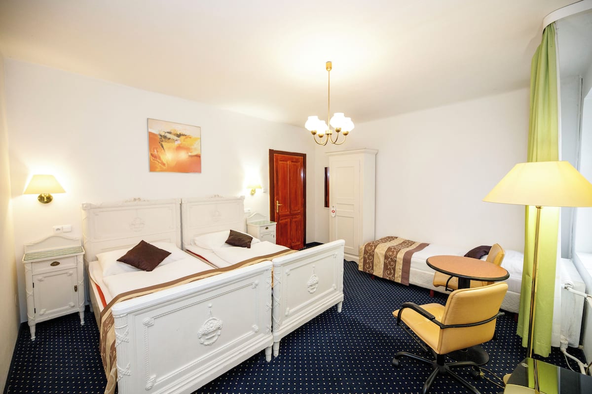 Habitación de hotel con cama doble, cama individual, escritorio con silla, lámpara y un cuadro en la pared.