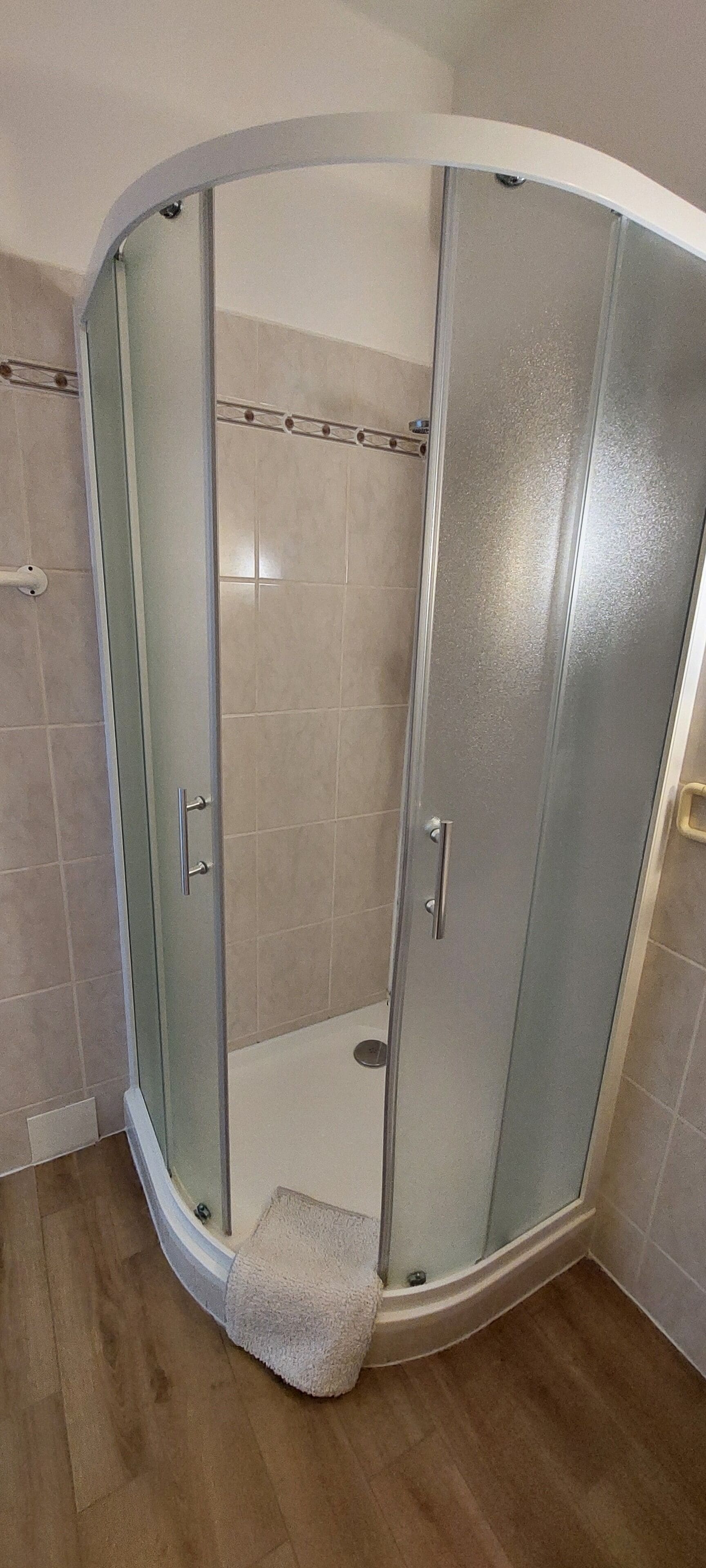 A corner shower with glass doors, a wooden floor, and beige tiles.