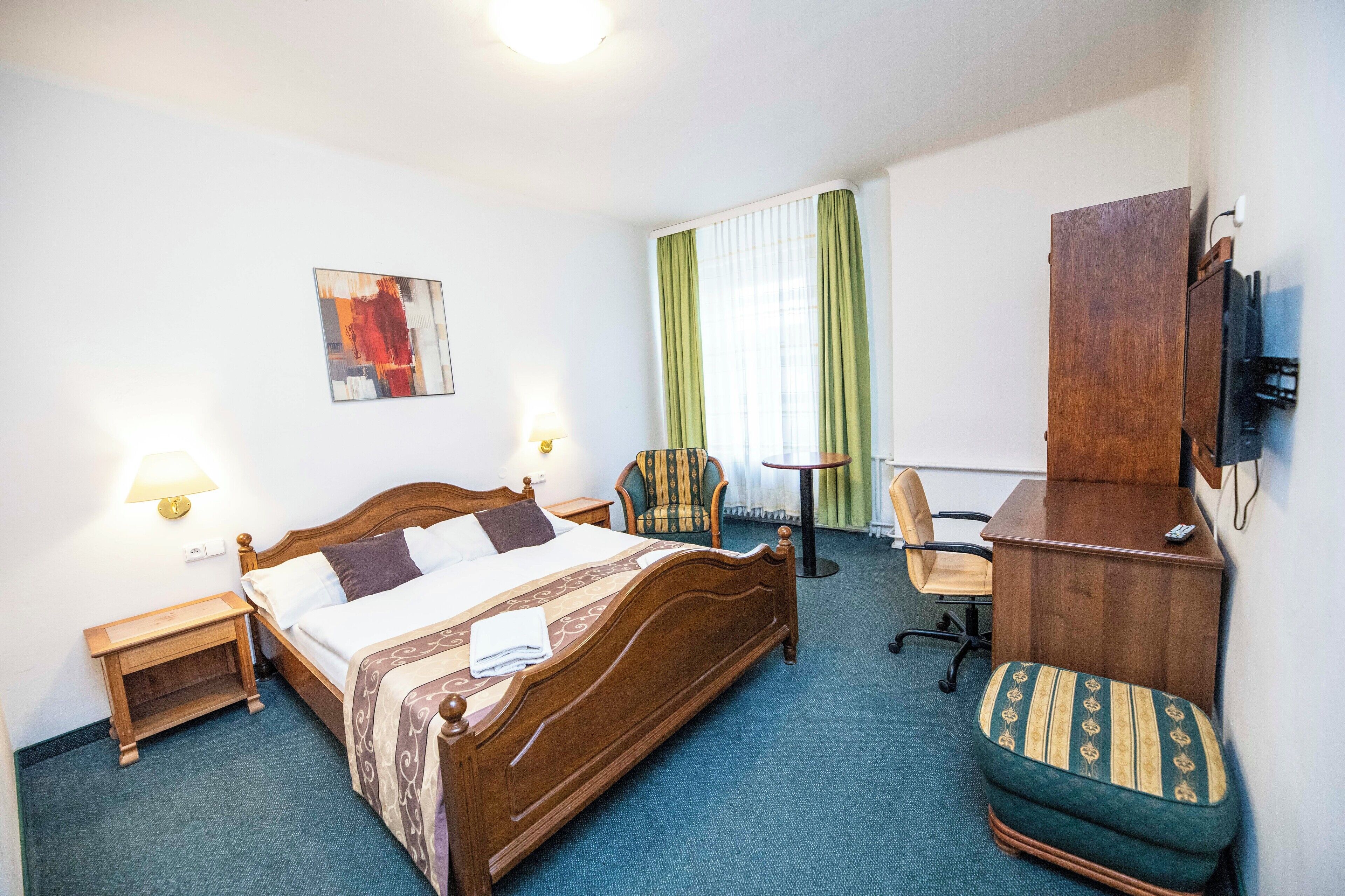 A hotel room with a wooden bed, a desk, a chair, a TV, and a painting on the wall.