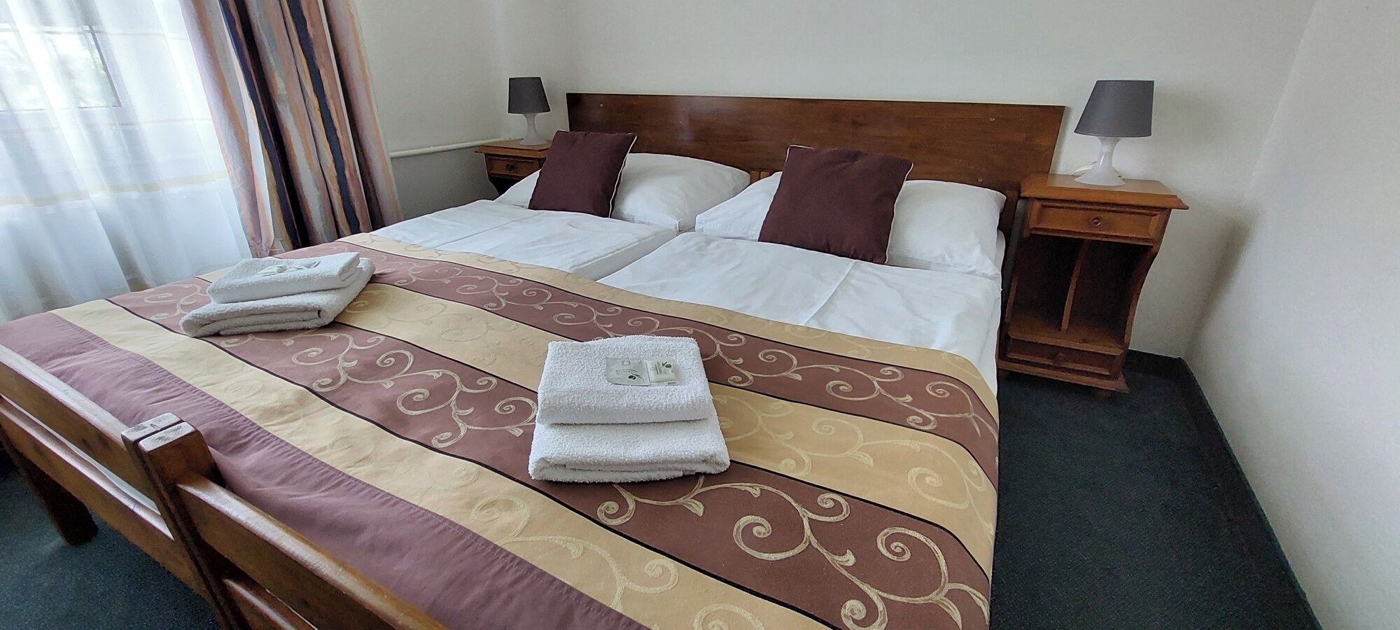 Ein ordentlich gemachtes Bett mit einem gemusterten Bettbezug, zwei Kissen und zwei gefalteten Handtüchern. Zwei Nachttische mit Lampen und ein Holzbeistelltisch sind ebenfalls vorhanden.