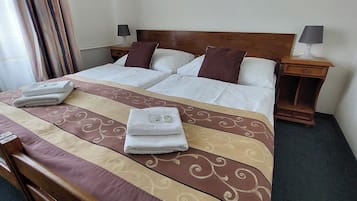 Un letto rifatto con cura, con una coperta a motivi, due cuscini e due asciugamani piegati. Sono presenti due comodini con lampade e un piccolo tavolo di legno.