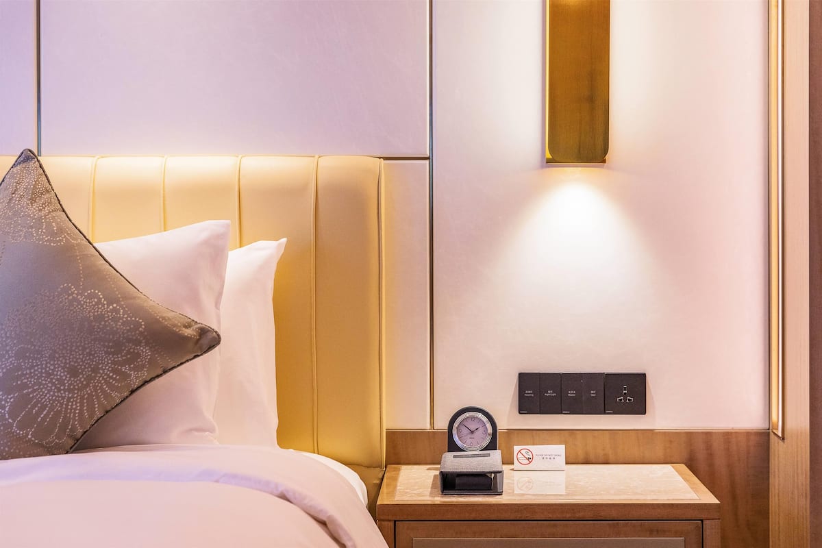 A hotel room with a bed, a nightstand, a clock, and a control panel.