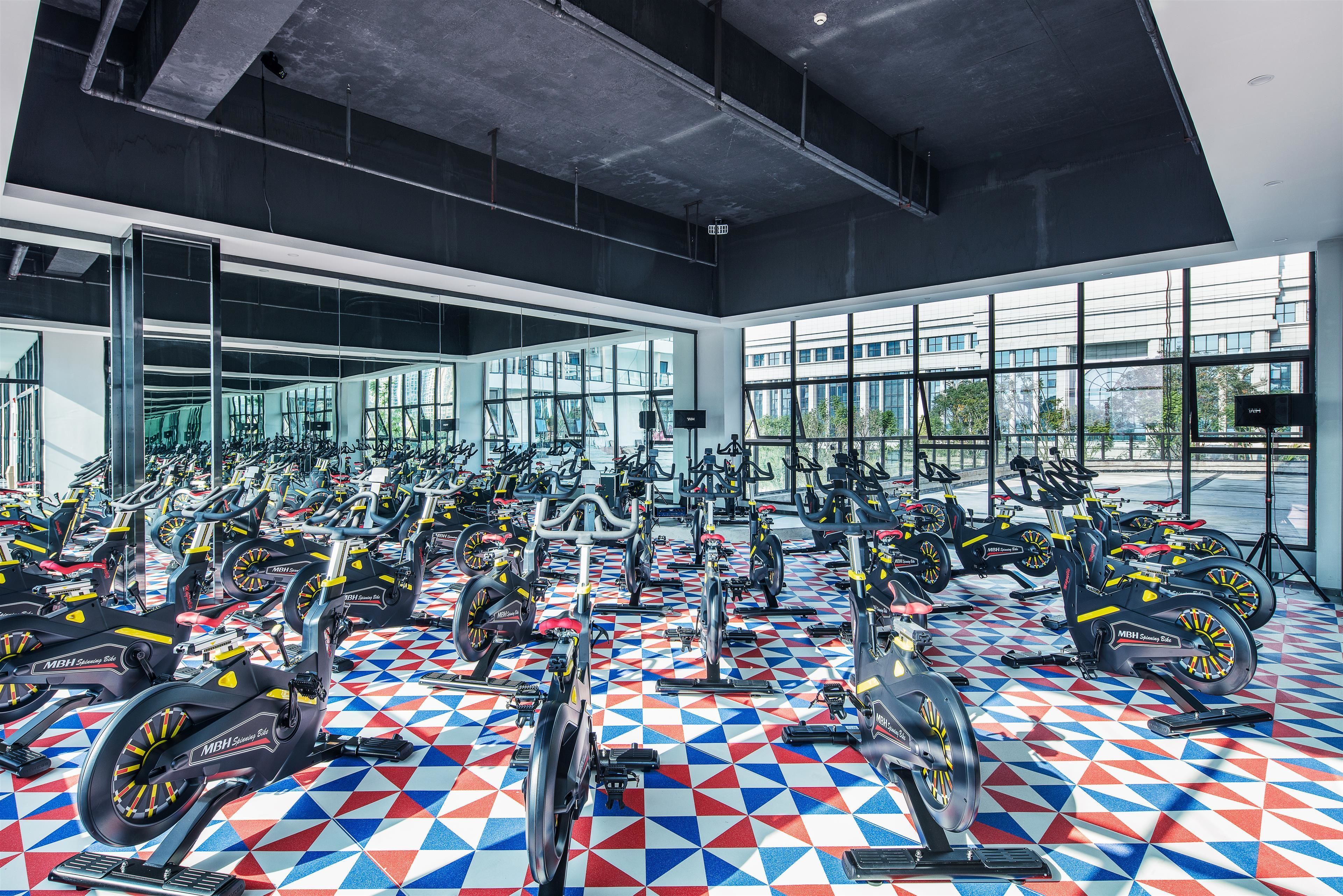Ein Fitnessraum mit mehreren in Reihen angeordneten Ergometer, großen Fenstern und einem geometrisch gemusterten Boden.
