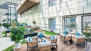 Un patio interior amplio con mobiliario de mimbre y plantas en macetas, ventanas grandes y vistas de la ciudad.