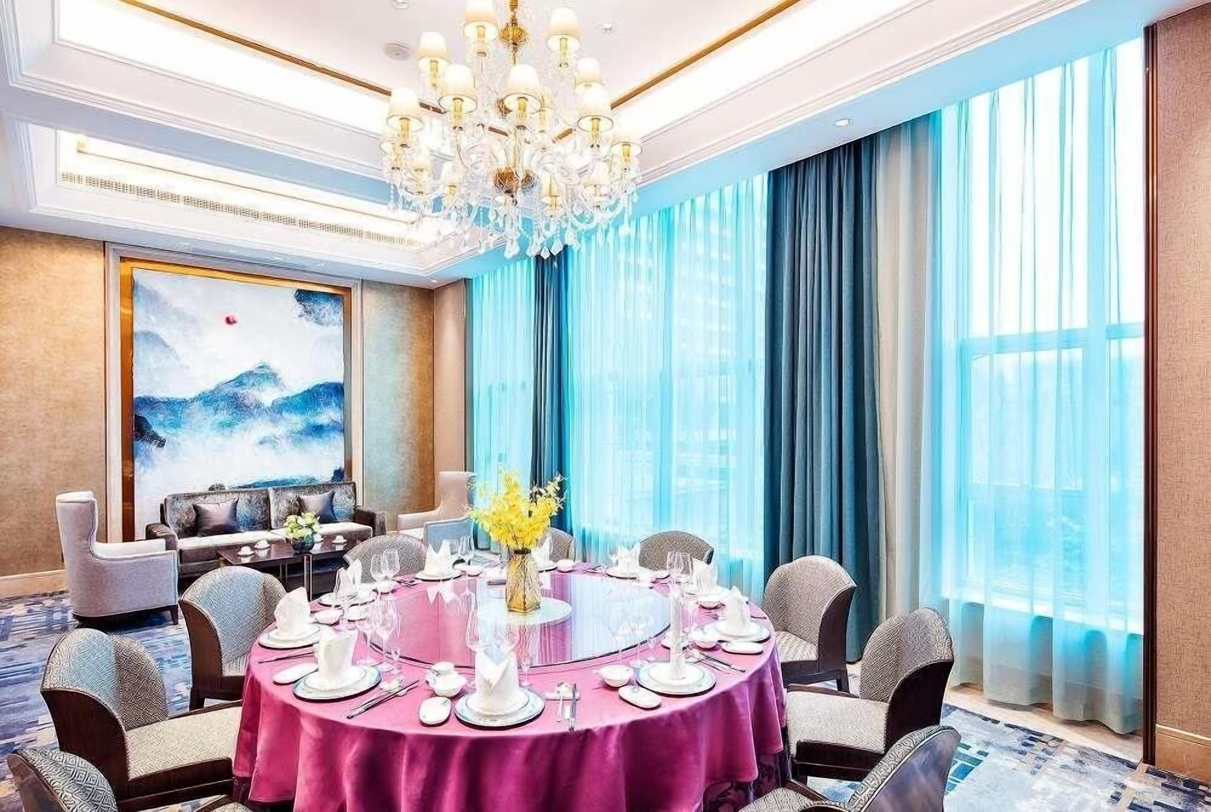 Comedor con una mesa redonda dispuesta para una comida, una araña y un ventanal con cortinas azules.