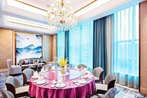 Comedor con una mesa redonda dispuesta para una comida, una araña y un ventanal con cortinas azules.