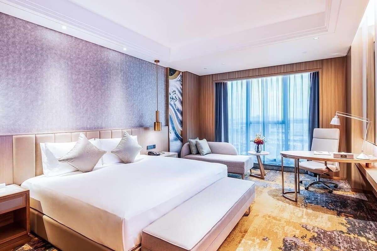 A modern hotel room with a large bed, a sofa, a desk, and a view of the city.