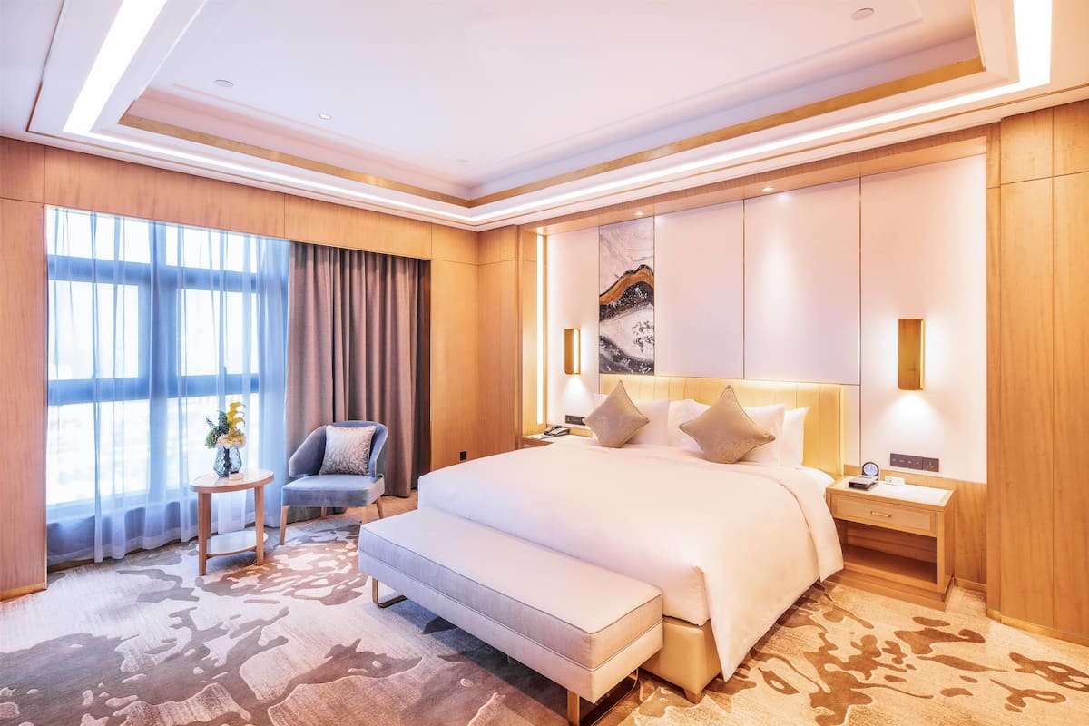 A spacious hotel room with a large bed, a bedside table, a chair, and a view of the city through large windows.