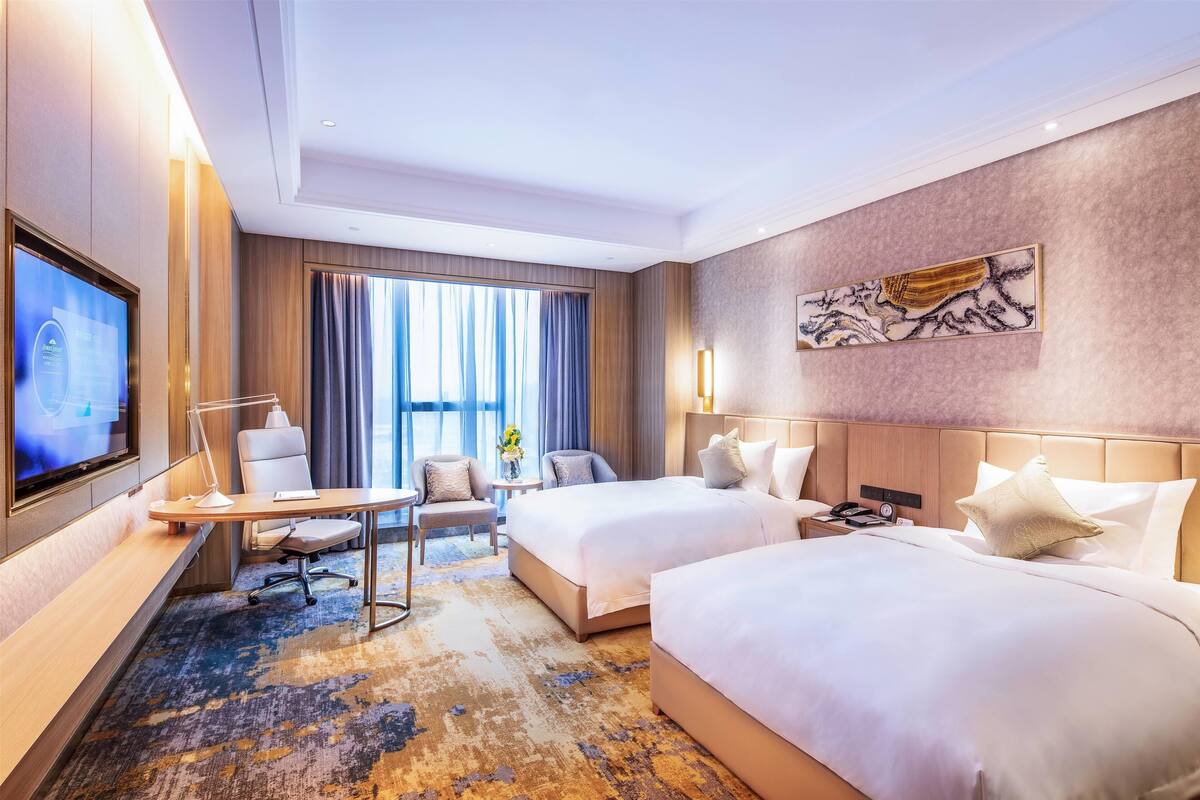 A hotel room with two beds, a desk, a TV, and a large window with curtains.