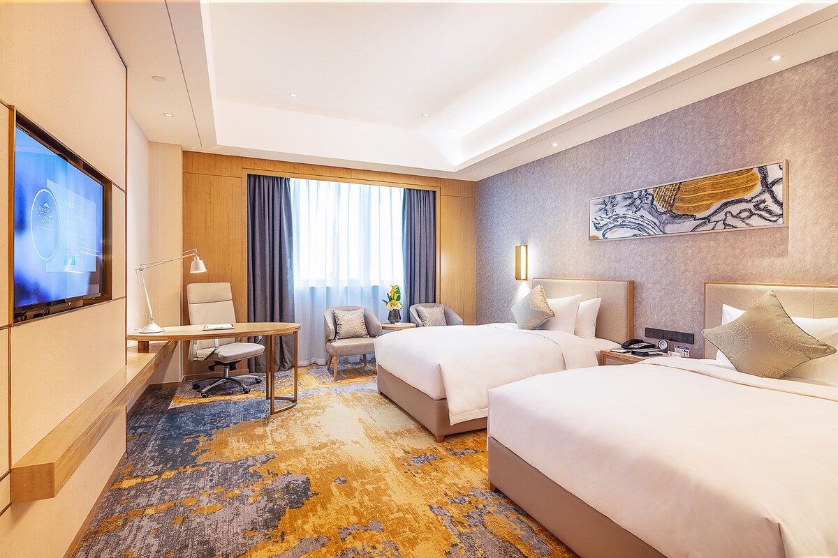 Ein Hotelzimmer mit zwei Betten, einem Schreibtisch, einem Sessel, einem Fernseher und einem großen Fenster mit Vorhängen.