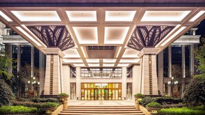 Una entrada de hotel moderna con una amplia fachada de vidrio, un acceso central con puertas dobles y un diseño simétrico con columnas y una estructura de techo geométrica.