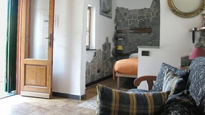 Una habitación acogedora con puerta de madera, pared de piedra, cama con manta naranja, sofá azul con estampado y un espejo redondo en la pared.