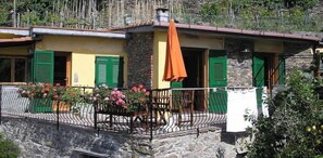 A stone building with green shutters, a balcony with a table and chairs, and a hanging orange cloth. - Camere Giuliano (Vernazza)