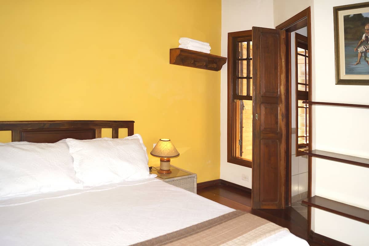 A bed with white linens, a nightstand with a lamp, a wooden shelf with a towel, and a door leading to another room.