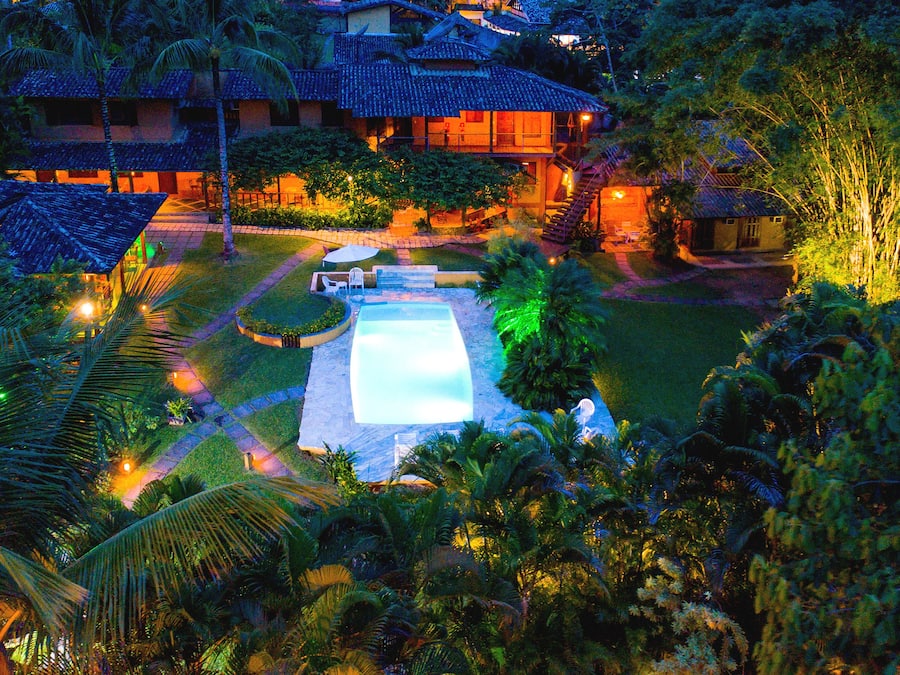 Una gran piscina bien iluminada rodeada de abundante vegetación y palmeras, con un edificio al fondo.