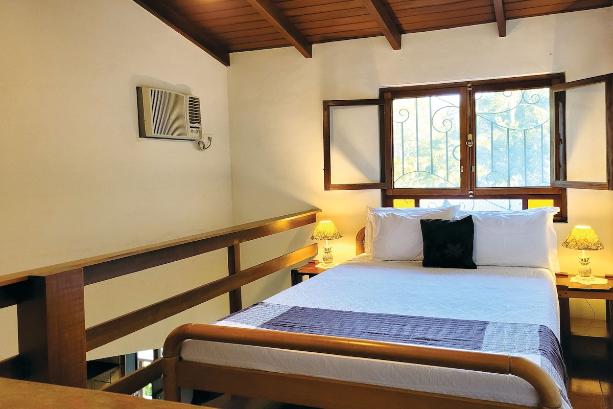 A bedroom with a wooden bed frame, white bedding, two bedside lamps, a window with decorative iron bars, and a wall-mounted air conditioning unit.