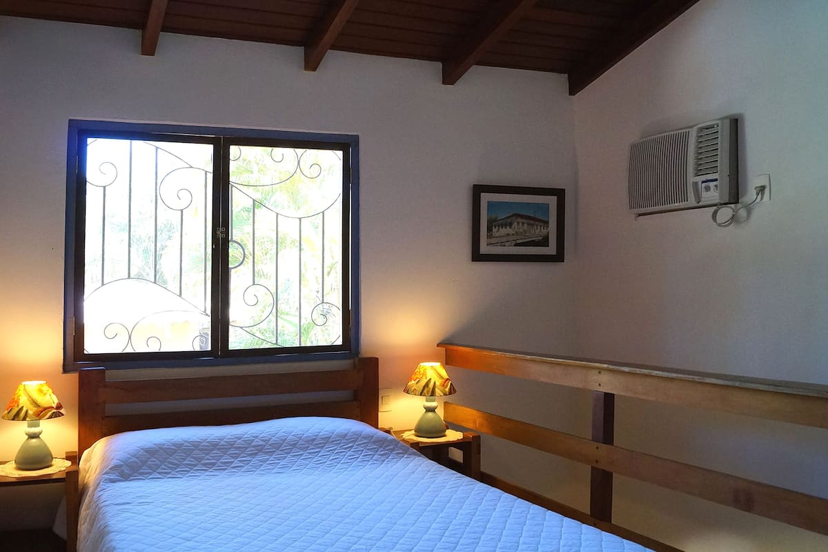 A bedroom with a bed, two bedside tables with lamps, a window with decorative iron bars, a wall-mounted air conditioner, and a framed picture.