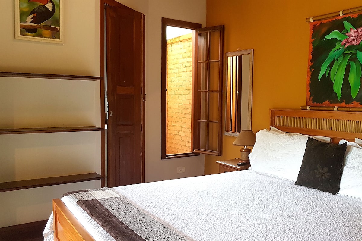 A bedroom with a bed, a nightstand, a lamp, a painting, and a door leading to a brick wall.