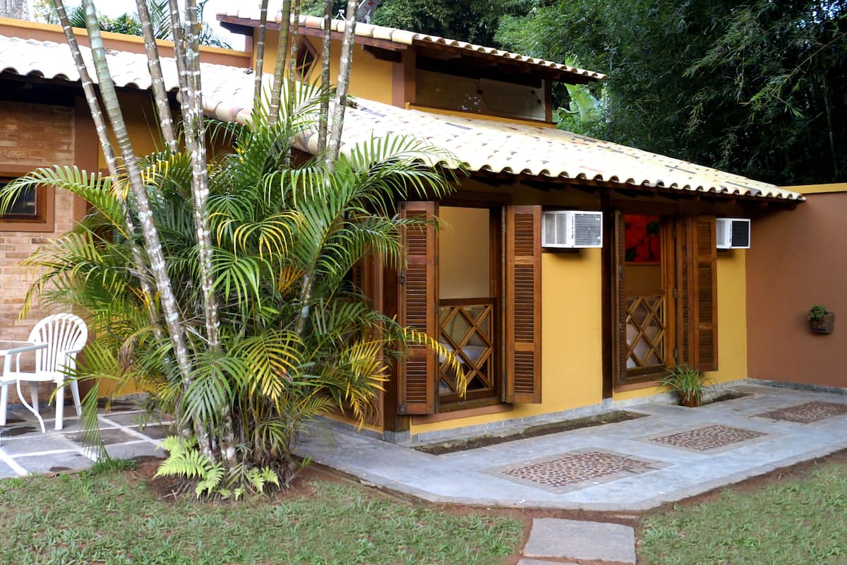 A two-story building with a yellow facade, wooden shutters, and a tiled roof. There is a small garden area with a white chair and palm trees.