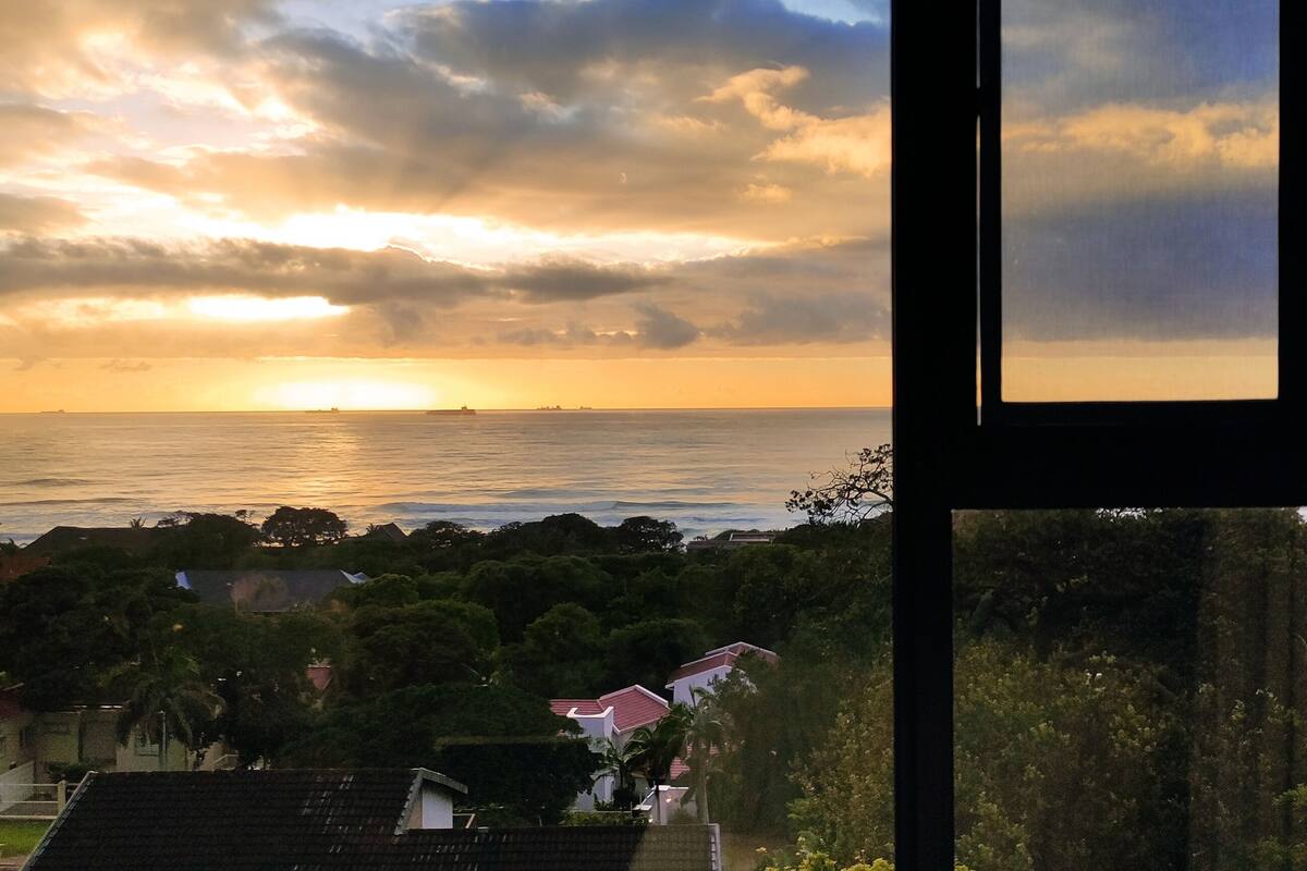 A coastal view from a window overlooking a sea with a sunset, houses, and greenery.