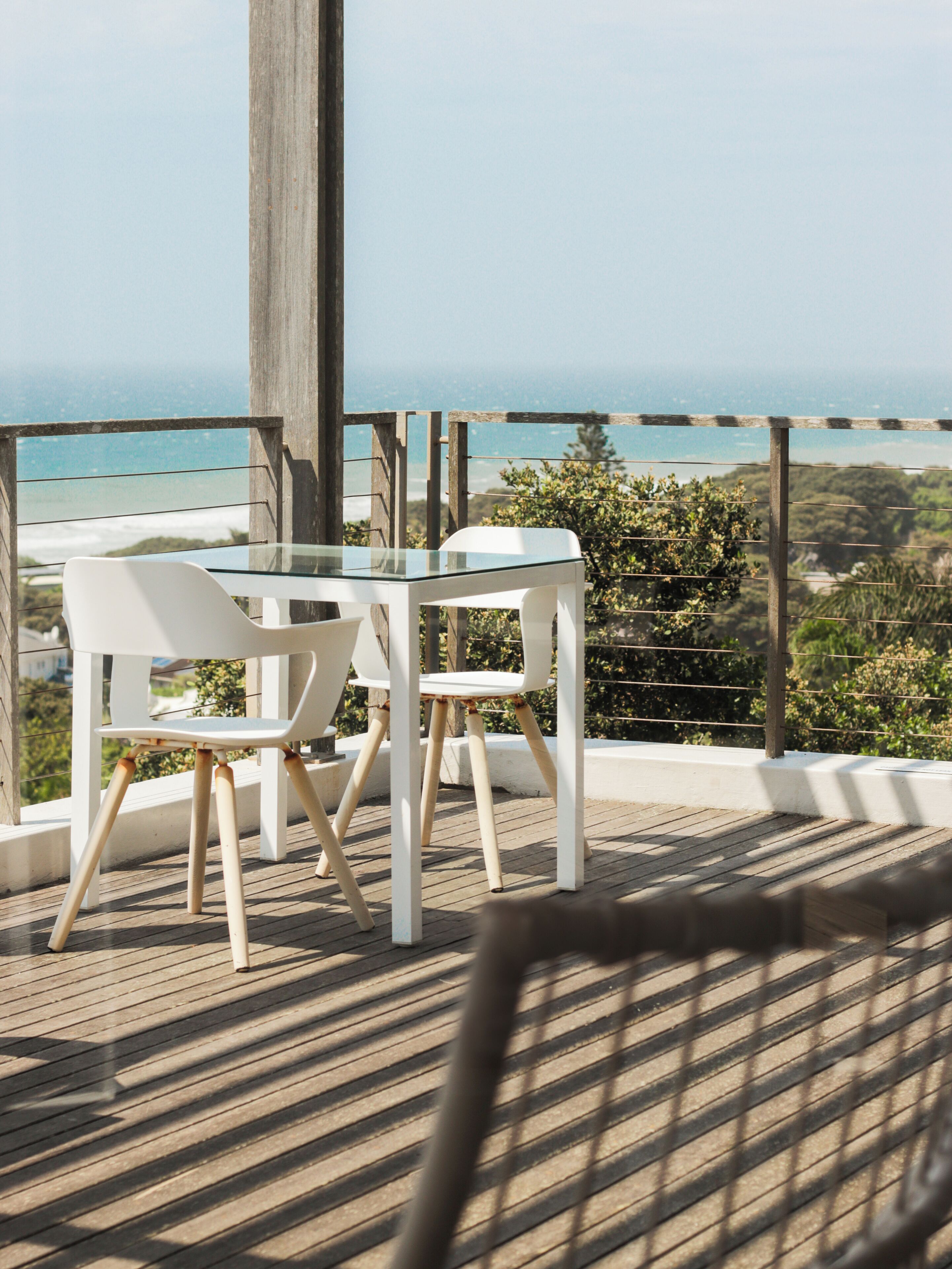 Un balcón con una mesa de vidrio y dos sillas blancas, con vistas a la costa.