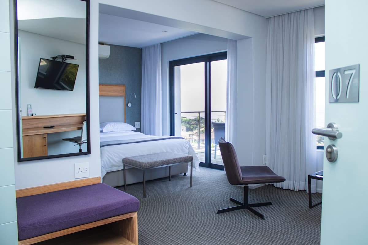 A hotel room with a bed, a bench, a chair, a TV mounted on the wall, and a balcony with curtains.
