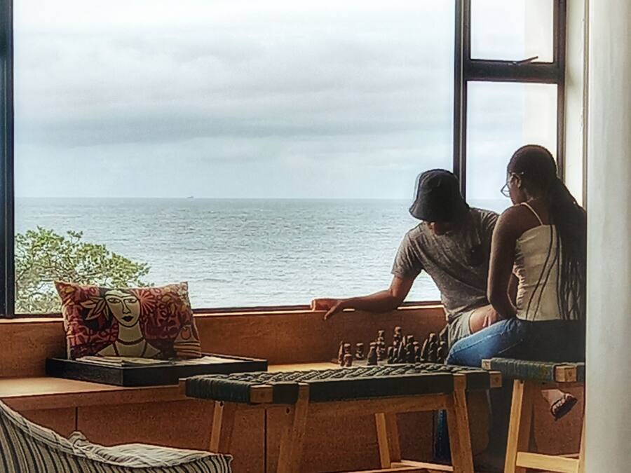 A room with a large window overlooking the sea, a wooden bench, and a chessboard.