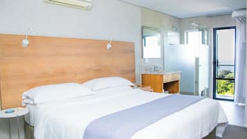 A hotel room with a large bed, a wooden headboard, a bedside table with a lamp, a bathroom with a mirror, and a view of greenery outside.