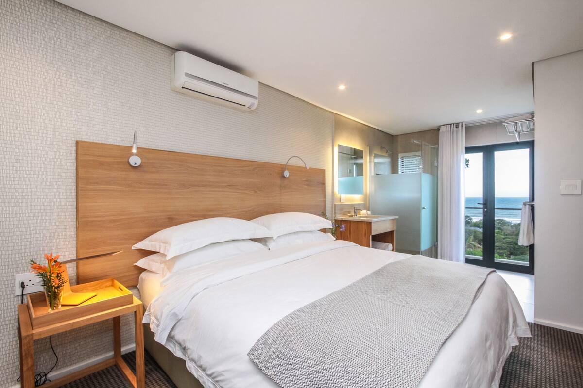 A hotel room with a large bed, a wooden headboard, a bedside table with a lamp, a mirror, and a view of the outdoors through a sliding door.