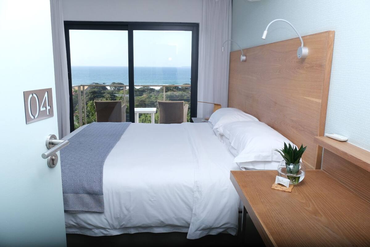A hotel room with a large bed, a wooden headboard, a bedside table with a plant, and a view of the ocean through a large window.