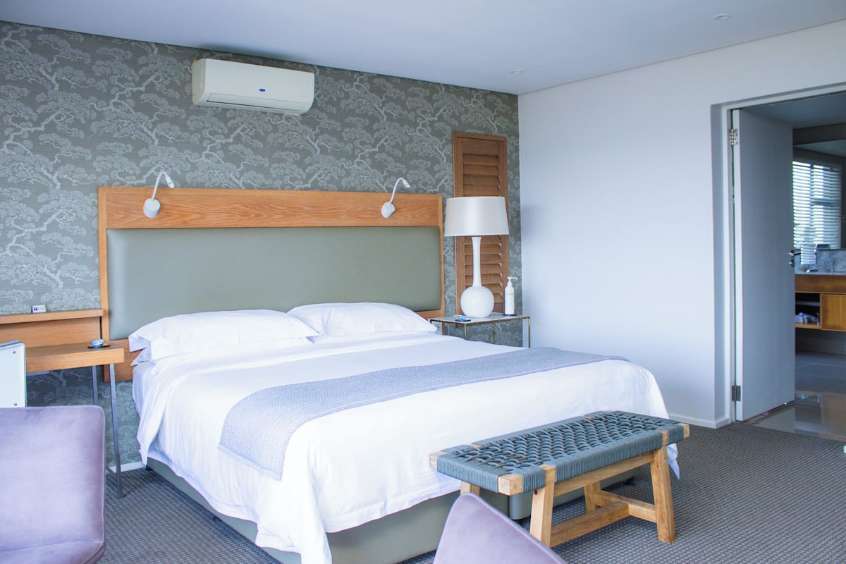A hotel room with a large bed, bedside tables, a bench, and a nightstand with a lamp.