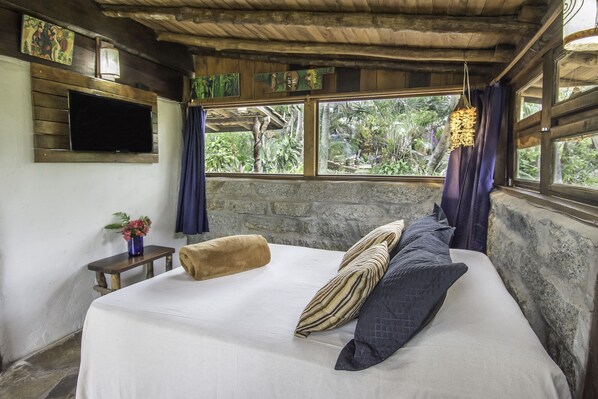 A bedroom with a bed, a TV mounted on the wall, a small table with a vase of flowers, and a view of greenery through the window. - Pousada Morro das Palmeiras (Imbituba)