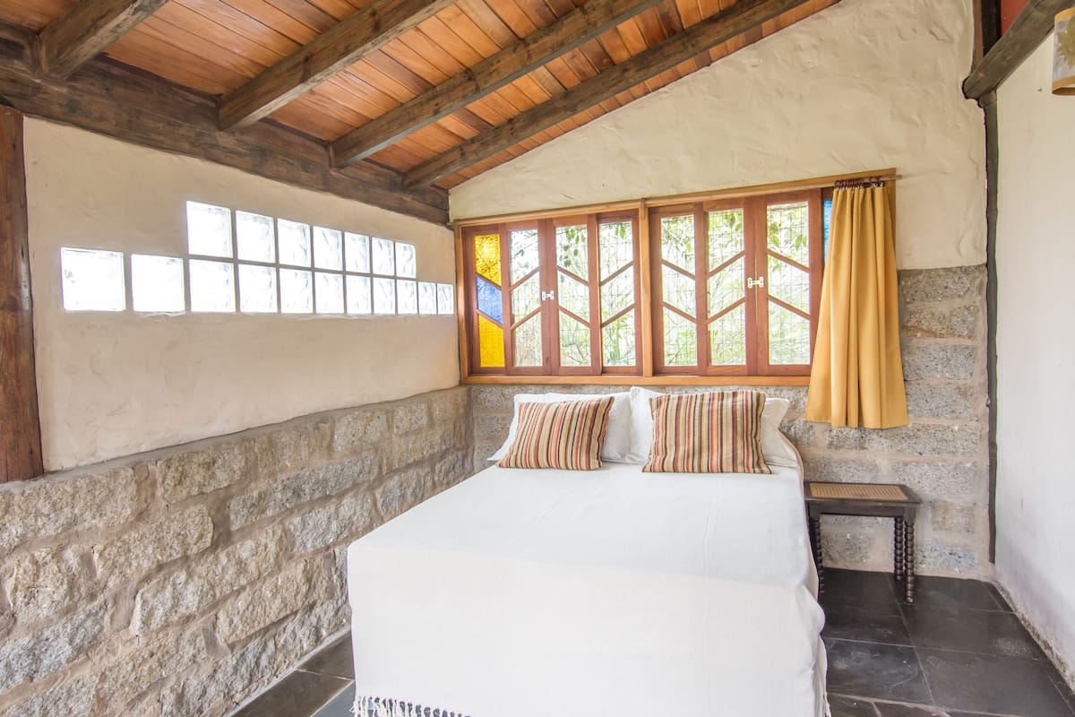 Ein Schlafzimmer mit Steinsandsteinwand, Holzbalken, einem Bett mit gestreiften Kissen, einem kleinen Tisch und einem Fenster mit gelbem Vorhang.