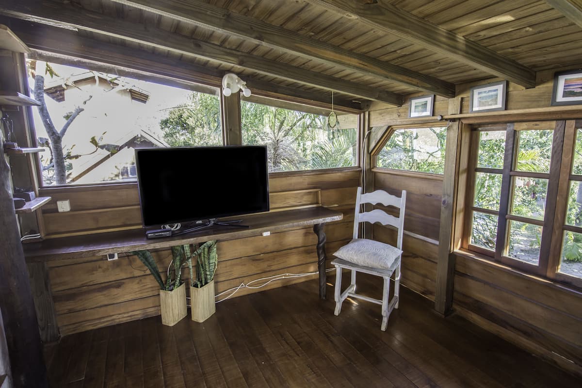 Ein Interieur einer Holzhütte mit einem Fernseher auf einem Regal, einem weißen Stuhl und großen Fenstern mit Blick auf Bäume.