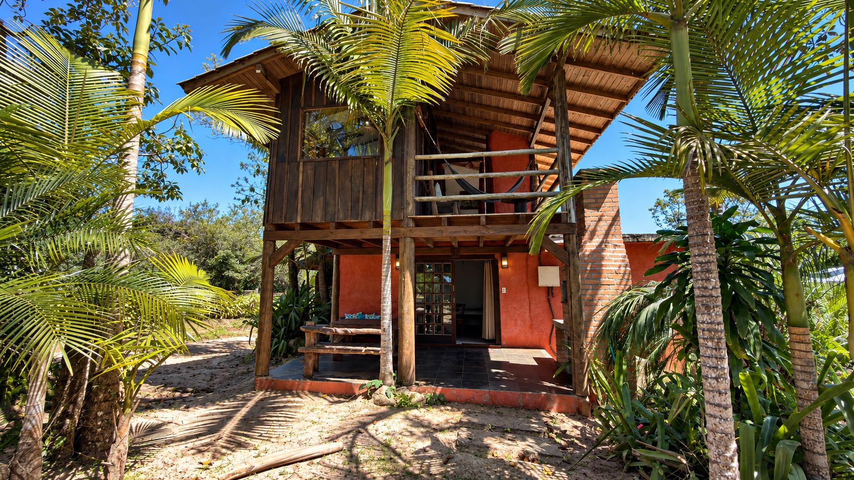 A two-story wooden structure with a balcony, surrounded by palm trees and a clear sky.